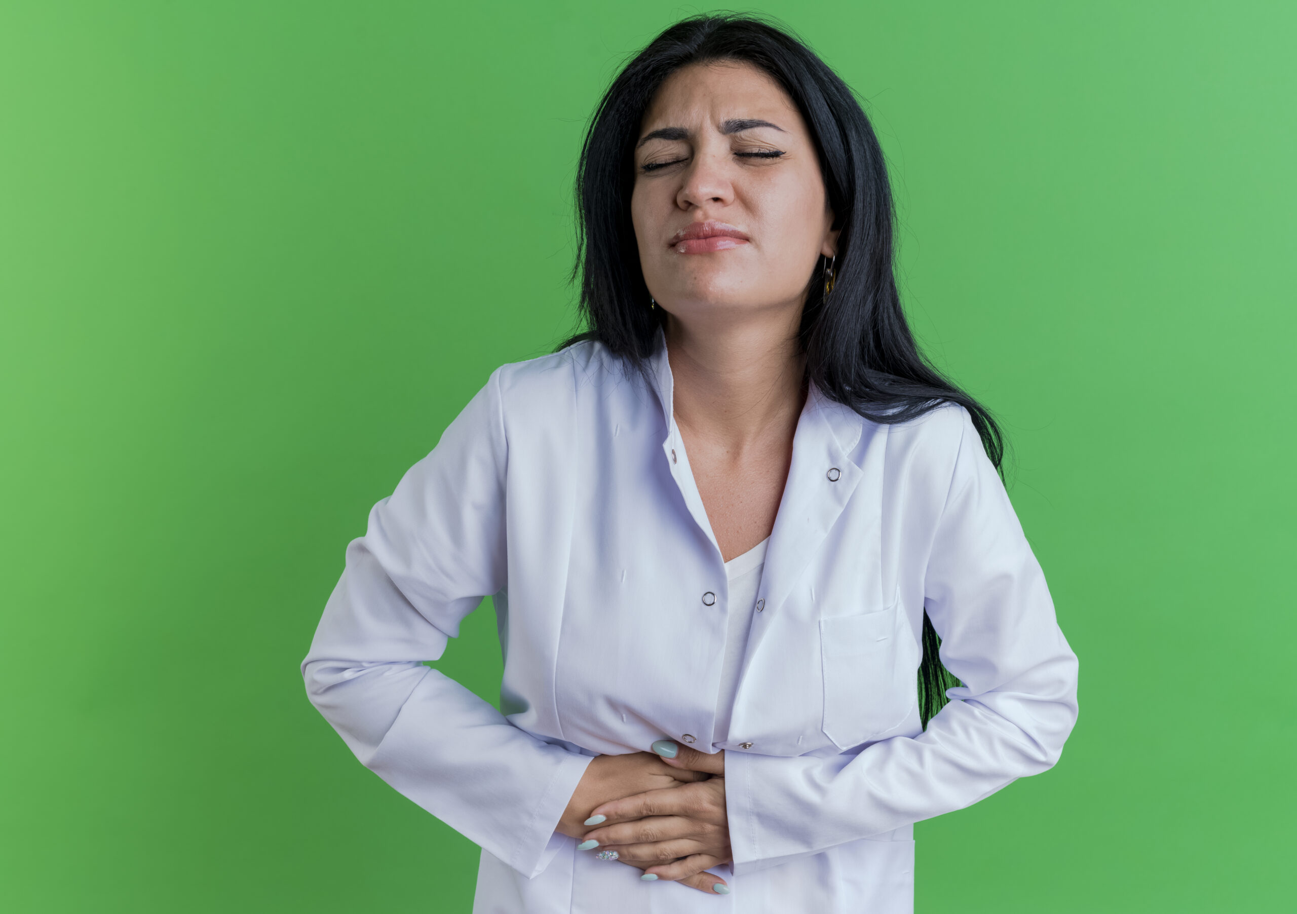 Aching young female doctor wearing medical robe holding belly with closed eyes isolated on green background with copy space